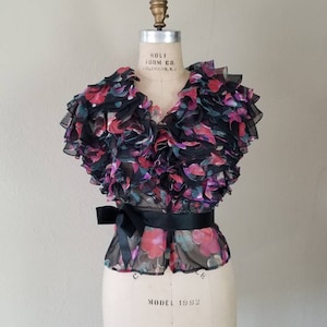May include: A vintage floral blouse with a ruffled collar and a black ribbon belt. The sheer fabric features a pattern of pink, purple, and green flowers on a black background. The blouse is displayed on a vintage dress form.