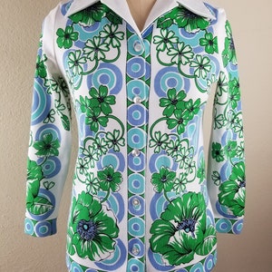 May include: A white button-down shirt with a vibrant floral pattern in green and blue. The shirt features a classic collar and long sleeves, with a series of buttons down the front. The floral design includes large flowers and circular patterns.