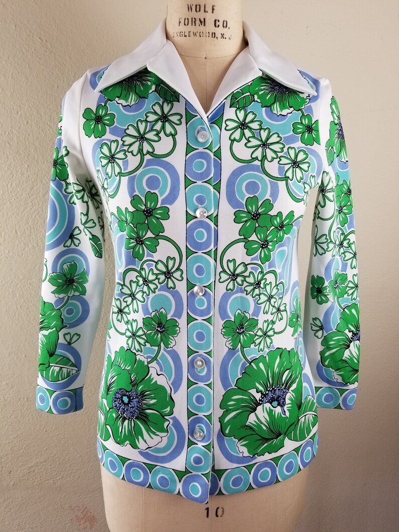 May include: A white button-down shirt with a vibrant floral pattern in green and blue. The shirt features a classic collar and long sleeves, with a series of buttons down the front. The floral design includes large flowers and circular patterns.