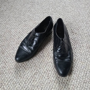 May include: A pair of black leather dress shoes with laces. The shoes have a classic design with a pointed toe and a low heel. The leather appears to be slightly worn, giving them a vintage look.