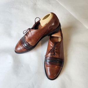 May include: A pair of brown leather dress shoes with a striped design across the vamp. The shoes have a classic lace-up closure and are displayed with wooden shoe trees inside. The shoes are on a white background.