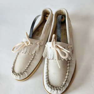 Puede incluir: Un par de mocasines de cuero blanco con detalles de flecos y un ribete trenzado. Los zapatos tienen una suela de color crema y una etiqueta de cuero marr&oacute;n que dice "Sebago Camp Moc".