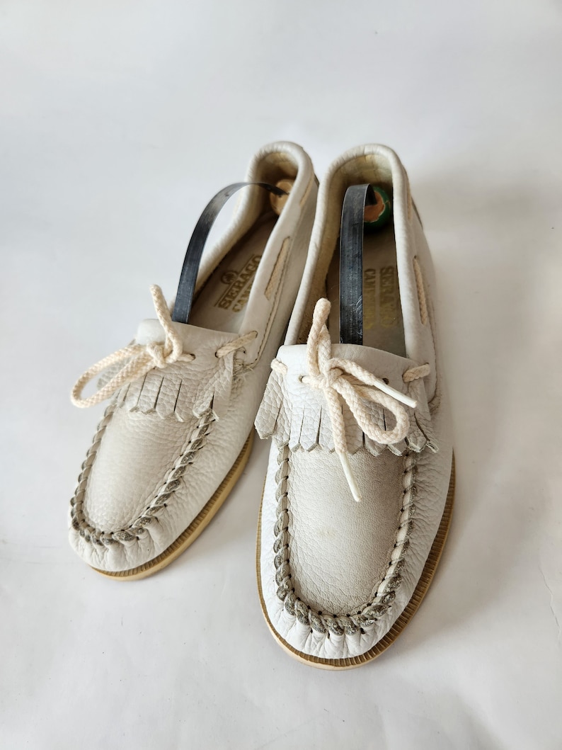 Puede incluir: Un par de mocasines de cuero blanco con detalles de flecos y un ribete trenzado. Los zapatos tienen una suela de color crema y una etiqueta de cuero marr&oacute;n que dice "Sebago Camp Moc".