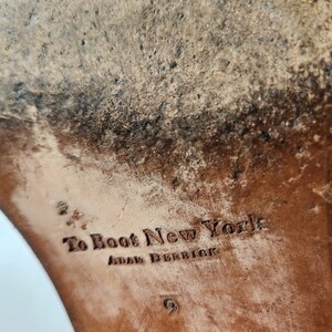 May include: Close-up of the sole of a brown leather boot with the text "To Boot New York" and "Adam Derrick" embossed on the leather.