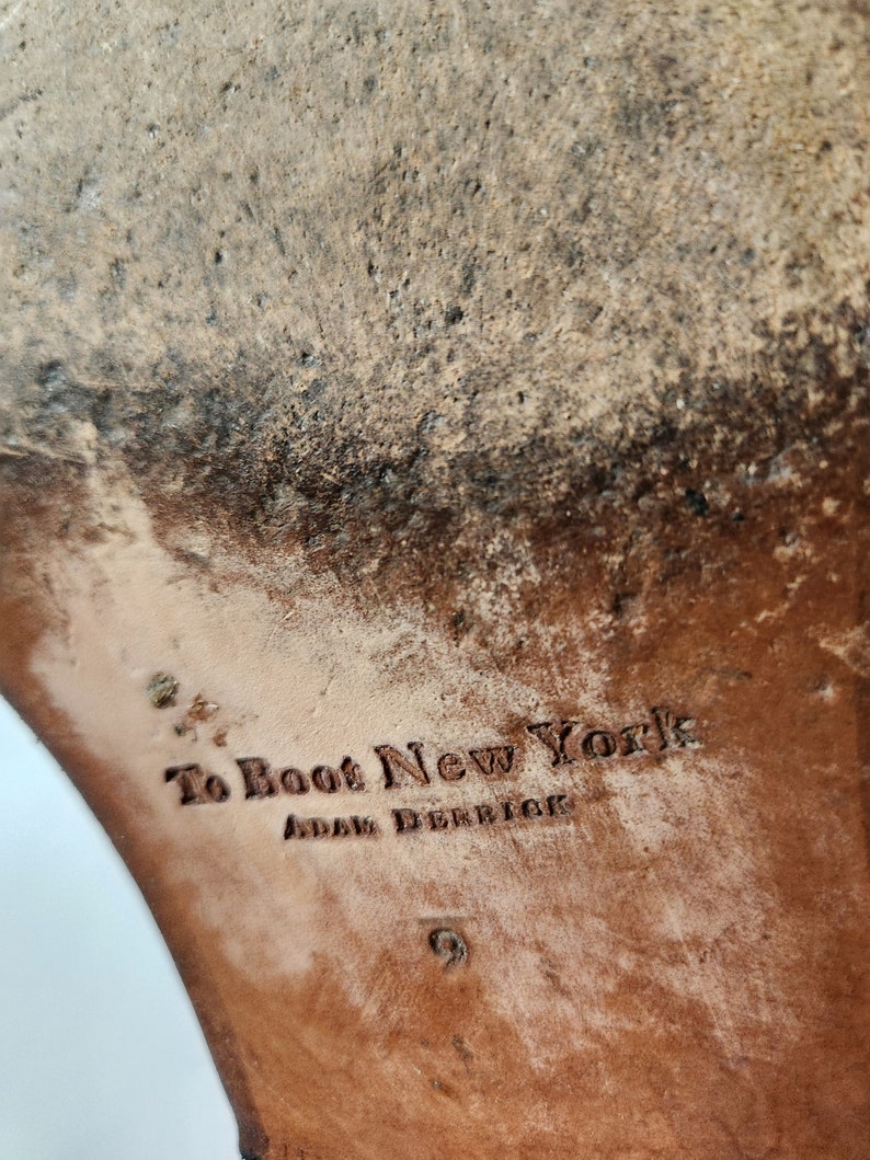 May include: Close-up of the sole of a brown leather boot with the text "To Boot New York" and "Adam Derrick" embossed on the leather.