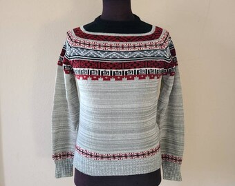 May include: A gray sweater with a black collar and red, white, and black geometric patterns across the chest and cuffs. The sweater is displayed on a mannequin and has long sleeves.