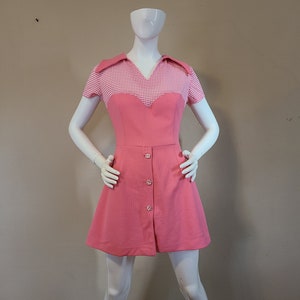 May include: A vintage pink dress with a white and pink gingham bodice and short sleeves. The dress features a V-neck collar, a fitted waist, and a button-down front. The skirt is A-line.