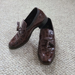 May include: A pair of brown leather loafers with a crocodile skin texture. The shoes have a tassel detail and are set on a neutral-colored carpet.