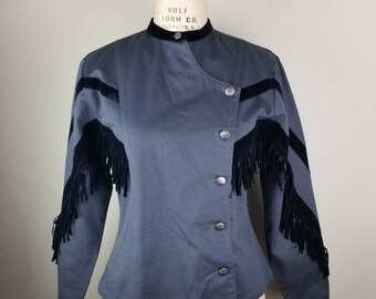 May include: A gray jacket with black fringe and accents. The jacket has a high collar, three silver buttons, and black fringe detailing on the sleeves and chest. The jacket is displayed on a mannequin.