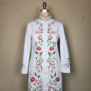 May include: A white, long-sleeved coat with a mandarin collar and floral embroidery. The embroidery features red and pink flowers with green leaves, creating a vertical design down the front and on the sleeves. The coat has a button-up closure.