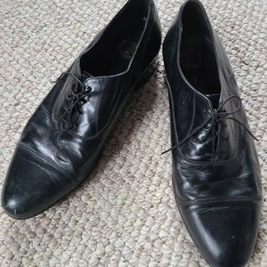 May include: A pair of black leather dress shoes with a classic Oxford design. The shoes have a pointed toe, lace-up closure, and a low heel. The leather appears to be polished and has a slight sheen. The shoes are displayed on a textured, neutral-colored surface.