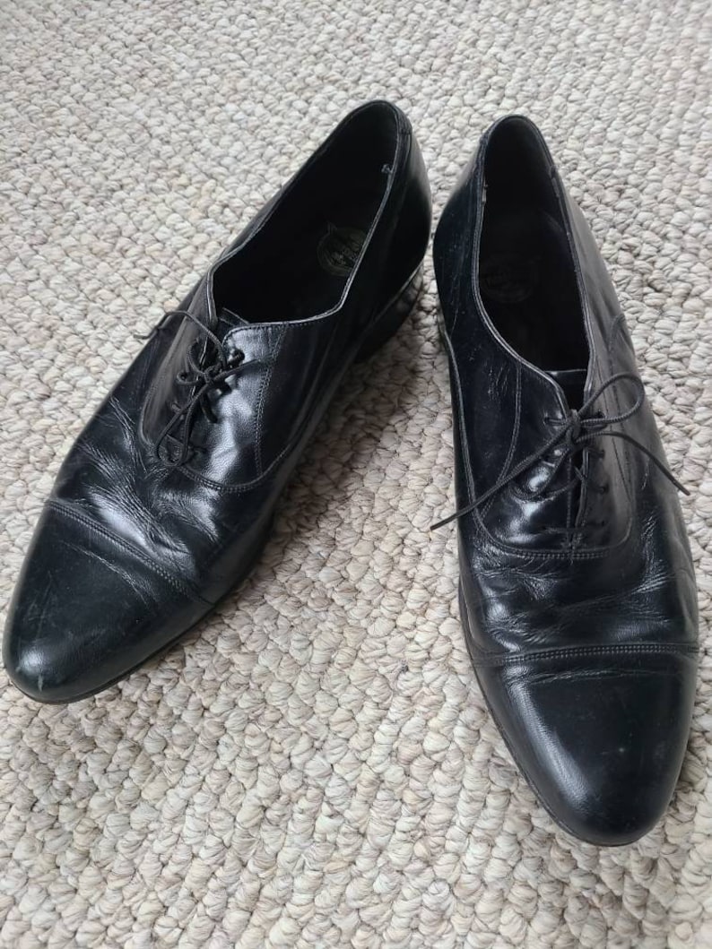 May include: A pair of black leather dress shoes with a classic Oxford design. The shoes have a pointed toe, lace-up closure, and a low heel. The leather appears to be polished and has a slight sheen. The shoes are displayed on a textured, neutral-colored surface.