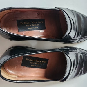 May include: A pair of black leather loafers with a slip-on design. The shoes have a label inside that reads "To Boot New York Adam Derrick Made in Italy."