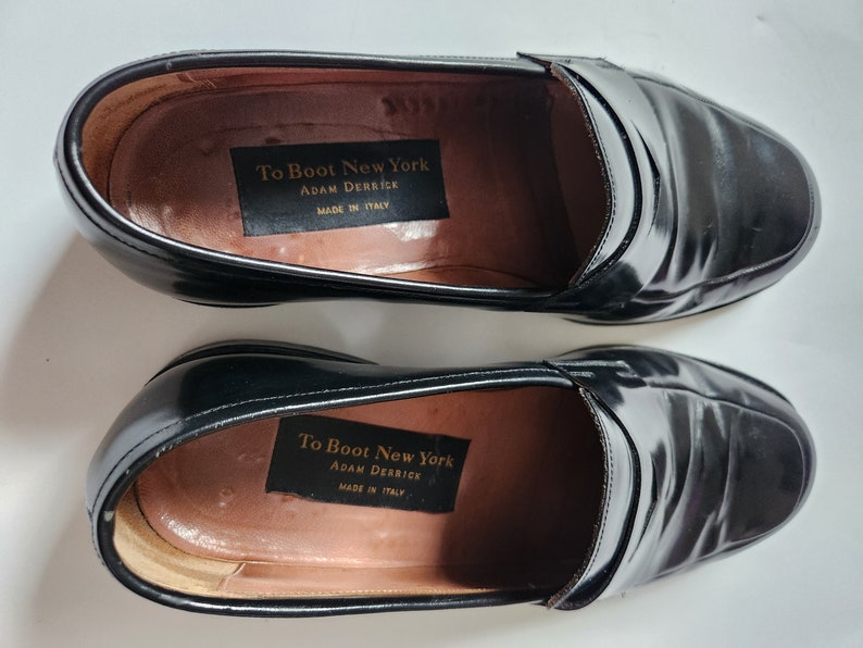 May include: A pair of black leather loafers with a slip-on design. The shoes have a label inside that reads "To Boot New York Adam Derrick Made in Italy."