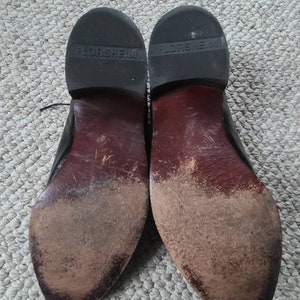 May include: Pair of well-worn black leather dress shoes, soles facing up. The soles are dark brown with visible wear and dirt. The brand name "FLORSHEIM" is embossed on the heel of each shoe.