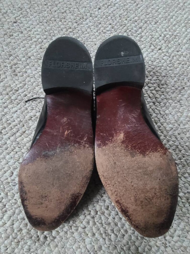 May include: Pair of well-worn black leather dress shoes, soles facing up. The soles are dark brown with visible wear and dirt. The brand name "FLORSHEIM" is embossed on the heel of each shoe.
