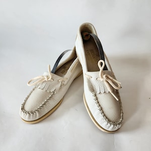 Puede incluir: Un par de zapatos n&aacute;uticos de cuero blanco con un detalle de flecos y un cord&oacute;n de algod&oacute;n blanco. Los zapatos tienen una suela de cuero marr&oacute;n y una etiqueta dorada que dice "Sebago Docksides" en el lateral.