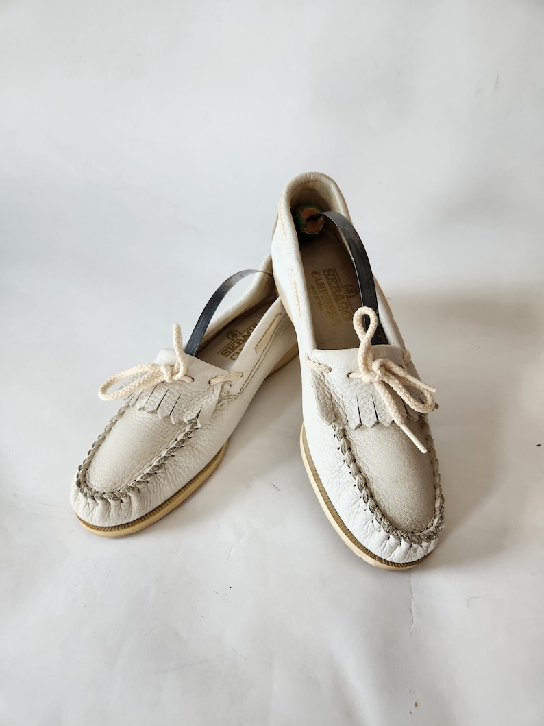 Puede incluir: Un par de zapatos n&aacute;uticos de cuero blanco con un detalle de flecos y un cord&oacute;n de algod&oacute;n blanco. Los zapatos tienen una suela de cuero marr&oacute;n y una etiqueta dorada que dice "Sebago Docksides" en el lateral.
