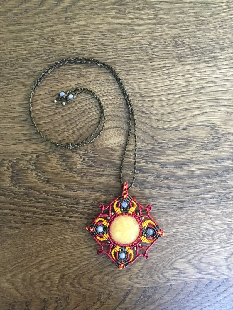 Puede incluir: Un collar con un colgante cuadrado con una piedra amarilla en el centro. El colgante est&aacute; hecho de cuerda roja, amarilla y azul y tiene un cord&oacute;n marr&oacute;n.