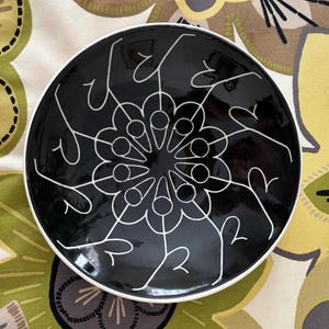 May include: Black and white ceramic plate with a geometric design. The plate features a circular pattern of white lines and dots on a black background.