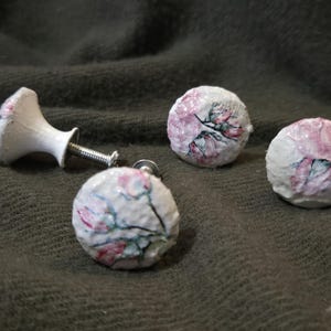 May include: A set of four decorative cabinet knobs. Three round knobs and one handle-style knob are visible. The knobs feature a white base with a floral design in pink and green. The hardware is silver.