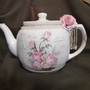 May include: A white ceramic teapot decorated with a pink rose floral print, cursive text, and a textured lace-like pattern. The lid and base are accented with white pearl trim. A single pink fabric rose and hanging crystal beads adorn the handle.