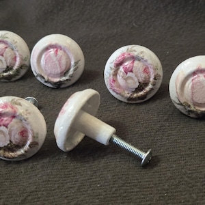 May include: Set of six white ceramic cabinet knobs with a floral design in pink and green. The knobs are round with a slightly domed top and a metal screw for attachment. The floral pattern is centered on each knob.