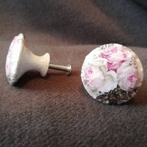 May include: Two white ceramic drawer knobs with a floral design. The knobs feature pink roses and green leaves on a white background.