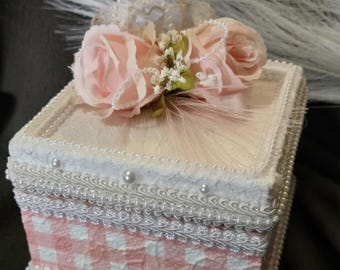 May include: A square decorative box featuring a pink and white gingham pattern on the sides, adorned with white lace trim and pearl accents. The lid is topped with two artificial pale pink roses, small white flowers, and pearl strands.