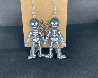 Puede incluir: Un par de pendientes de Halloween con figuras de esqueletos. Cada pendiente tiene una cabeza negra con rasgos faciales blancos y un cuerpo de esqueleto blanco. Los pendientes cuelgan de ganchos plateados y se exhiben en una tarjeta marrón.