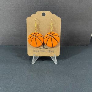 May include: A pair of orange and black basketball-themed dangle earrings with gold hooks. The earrings are displayed on a brown card with the text "Sleepy Hollow Designs".