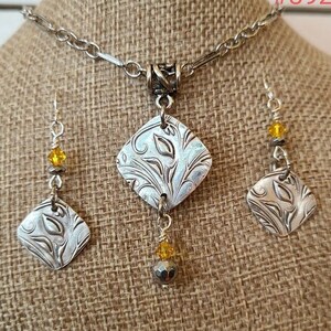 Peut inclure: Un ensemble de collier et boucles d'oreilles en argent. Le collier présente une chaîne et un pendentif en forme de losange avec un motif floral. Les boucles d'oreilles reprennent le motif du pendentif et comprennent des perles jaunes et grises. Les bijoux sont présentés sur une surface en toile de jute.