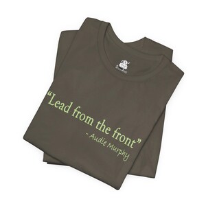 May include: Olive green t-shirt with the quote "Lead from the front" attributed to Audie Murphy in light green text. The shirt is folded and has a small logo on the upper right side.