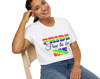 Op de afbeelding: Wit T-shirt met de tekst "PRIDE Free to be ME" in regenboogkleuren. Het shirt heeft een ronde hals en wordt gecombineerd met een blauwe spijkerbroek. De afbeelding toont een persoon die op een houten stoel zit.