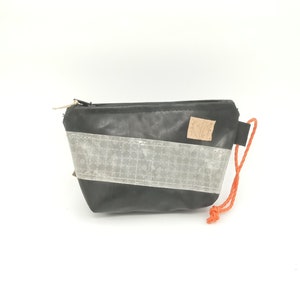 Könnte beinhalten: Kleine, schwarze Tasche mit diagonalem grauem Streifen und goldenem Reißverschluss. Eine orangefarbene Kordel ist an der Seite befestigt. Ein kleines, rechteckiges Korketikett befindet sich an der Seite der Tasche. Auf weißem Hintergrund.