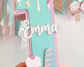 May include: A pastel pink, green, and purple layered cardboard number one with a white frosting design and sprinkles. The number one has the name "Emma" written on it. The number one is decorated with a pink and white ice cream cone, a pink and green popsicle, and a purple donut.