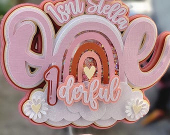 May include: A multi-layered cake topper featuring the text "Isn't Stella one-derful" in pink and white script. The design includes a central rainbow with glittery shaker elements, a white cloud base, and two small white flowers with yellow hearts.