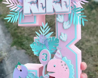 Könnte beinhalten: Eine Geburtstagdekoration aus Papier in Rosa, Blau und Lila. Das Design zeigt die Zahl 3, die Buchstaben "KK" und Dinosaurierfiguren. Dekorative Blätter und ein Ballon mit der Zahl 3 sind ebenfalls enthalten.