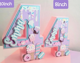 Op de afbeelding: Twee decoratieve cijfers 4, één van 25 cm hoog en de andere van 20 cm hoog, in pastelkleuren. De cijfers zijn lavendelkleurig met blauwe en roze accenten, versierd met ijs, snoep en de naam "Emma".