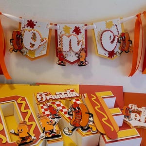 May include: A festive birthday banner and decorations with a hot dog theme. The banner spells out "one" with hot dog and condiment illustrations. The decorations include a pop-up display with the name "Franklin".
