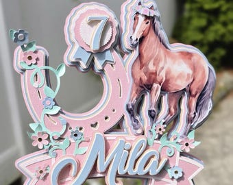 May include: A layered paper cake topper featuring a pink horseshoe, a brown horse, and a blue ribbon rosette with the number 7. The design includes small light blue and pink flowers on vines and a banner at the bottom with the name Mila in blue.