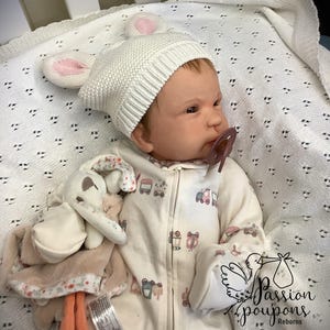 Può includere: Una bambola reborn indossa un cappello di lana bianco con orecchie da coniglio rosa e una tutina color crema con grafiche di veicoli. La bambola ha un ciuccio e tiene in mano un peluche. La bambola è sdraiata su una coperta bianca.
