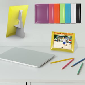 May include: A yellow picture frame with a stand, displaying a photo of a child running at a Sports Day 2026 event. The frame is shown in a horizontal orientation with a photo, and a vertical yellow frame with a stand is nearby, alongside colored pencils.