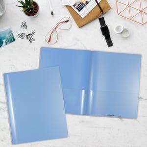 May include: Two light blue plastic folders, one shown closed and one shown open, featuring two interior pockets for document storage. The folders have a smooth, semi-gloss finish and are displayed on a white marble surface with office supplies.
