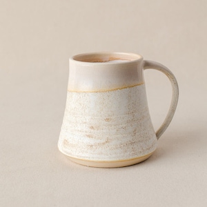 May include: A white ceramic mug with a beige rim and handle. The mug has a speckled glaze and a slightly flared shape.