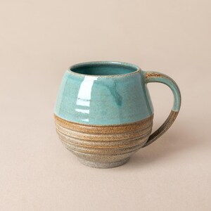 Peut inclure: Une tasse en céramique de forme arrondie avec une glaçure turquoise lisse et brillante sur la partie supérieure et une base brune texturée et mouchetée avec des stries horizontales. L'anse est finie dans le même ton brun moucheté.