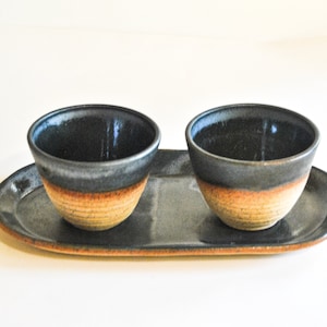 May include: Two small, dark blue ceramic bowls with a light brown band around the rim, sitting on a matching oval tray.