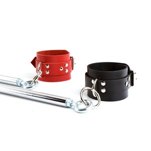 May include: A red leather cuff and a black leather cuff, each featuring silver-toned metal rivets and a large circular metal ring. The cuffs are connected to a long, cylindrical, chrome-finished metal spreader bar with swivel eye bolts.
