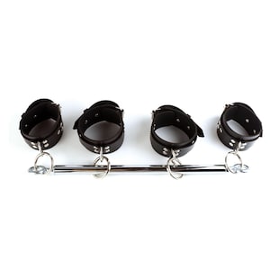 May include: A black leather spreader bar featuring four adjustable cuffs with silver-toned metal buckles and rivets. The cuffs are attached to a central polished chrome metal rod by four circular metal rings, allowing for movement along the bar.