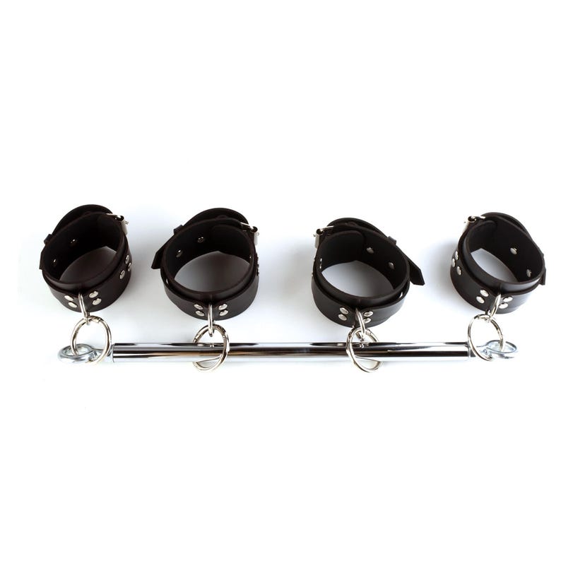 May include: A black leather spreader bar featuring four adjustable cuffs with silver-toned metal buckles and rivets. The cuffs are attached to a central polished chrome metal rod by four circular metal rings, allowing for movement along the bar.